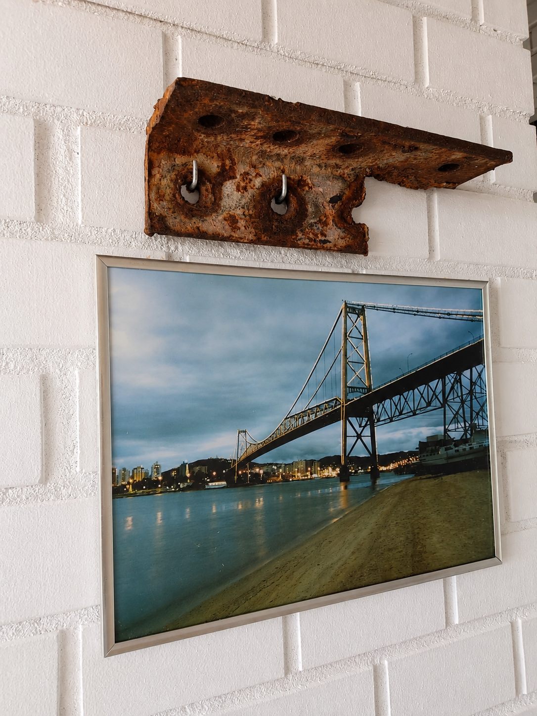 Fragmento real da Ponte Hercílio Luz preservado na parede de tijolinho da Lagoa Nômade, acima da foto emoldurada da ponte